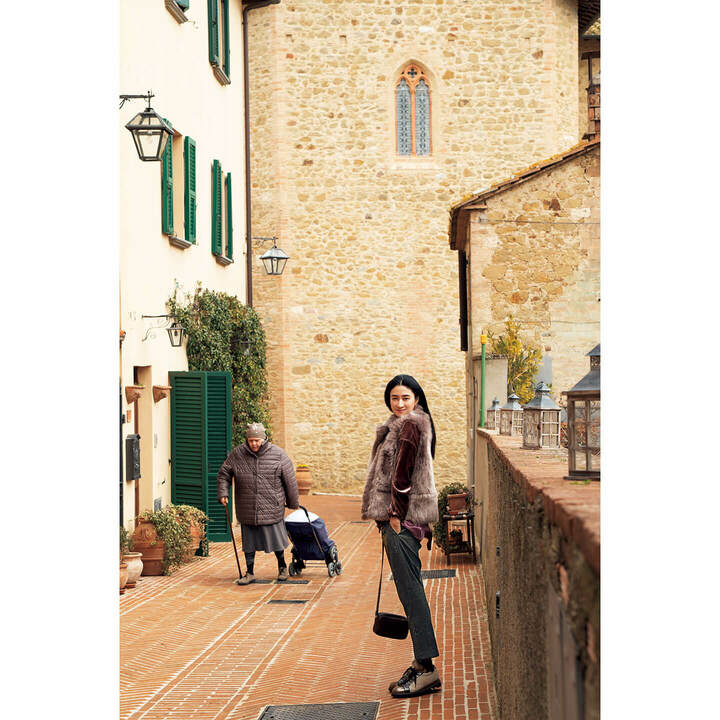 「この村の住人でしょうか？ 石畳の路地で出会ったおばあちゃまがかわいくて！ 彼女が被っていたニットキャップも、なんとクチネリだったんですよ」（小雪さん）。優しいグレージュ色と古い街並みが美しいハーモニーを醸し出します。ファーベスト￥969,000・ブルゾン［参考商品］・ブラウス［参考商品］・パンツ￥145,000・バッグ［参考商品］・靴［参考商品］（ブルネロ クチネリ ジャパン）