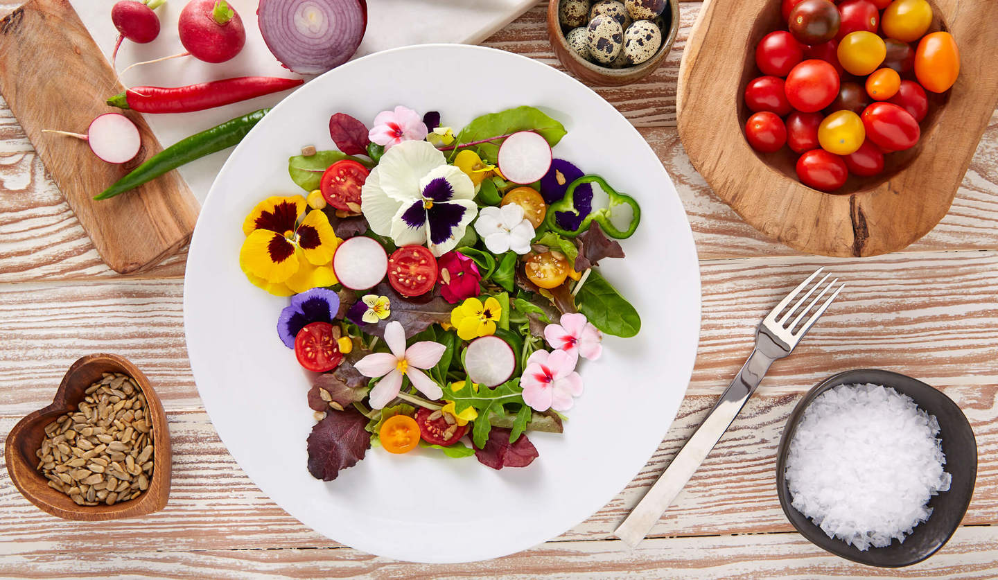 白いお皿の上に、パンジーなどの食べられるお花と赤と黄色のトマトで飾られた生野菜サラダがあり、そのお皿の周囲にフォークや塩、トマトなどが置かれている