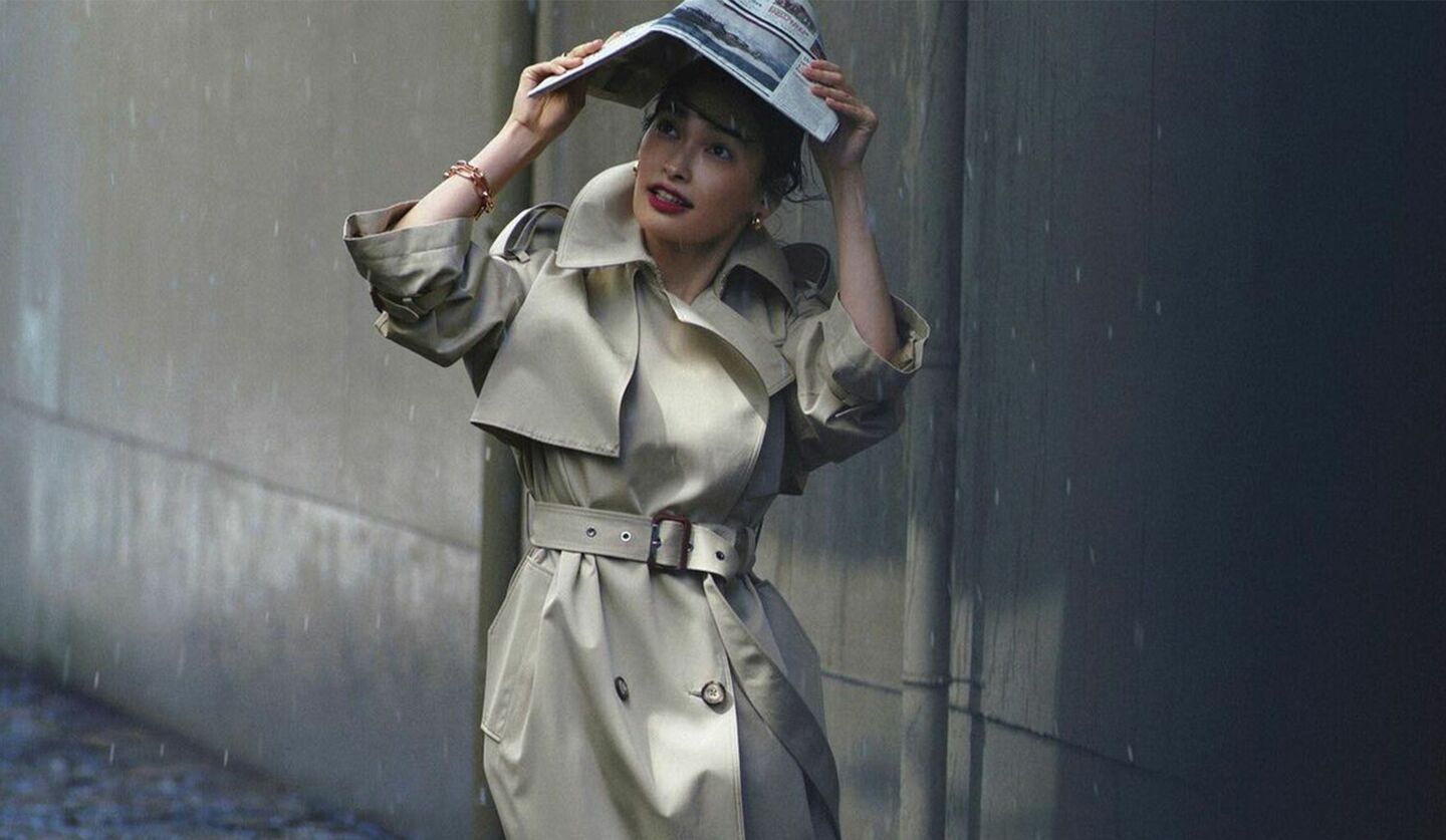 大政絢さんが新聞を傘代わりにしながらトレンチコートを着ている写真