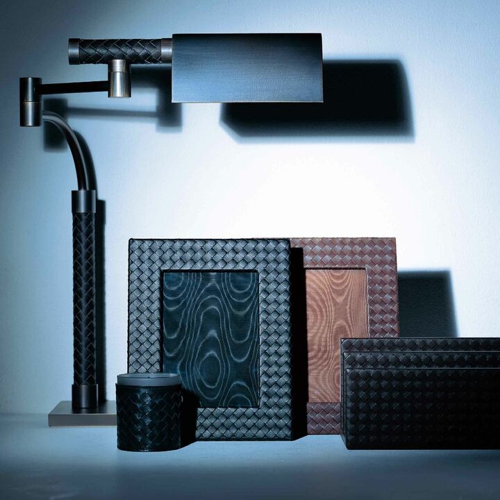 左から／リーディングランプは受注生産のため、ベースとなるフィニッシング（ブロンズ、ガンメタル、スチール）と、イントレチャートの色を革見本（６色）から選択。ヘッド部分に搭載されたタッチセンサーで、三段階調光が可能。¥460,000・フォトフレーム［縦24.7×横19.6㎝］各¥75,000・ウッディな香りに癒されるホームキャンドル¥25,000・レターホルダー¥114,000（ボッテガ・ヴェネタ ジャパン）