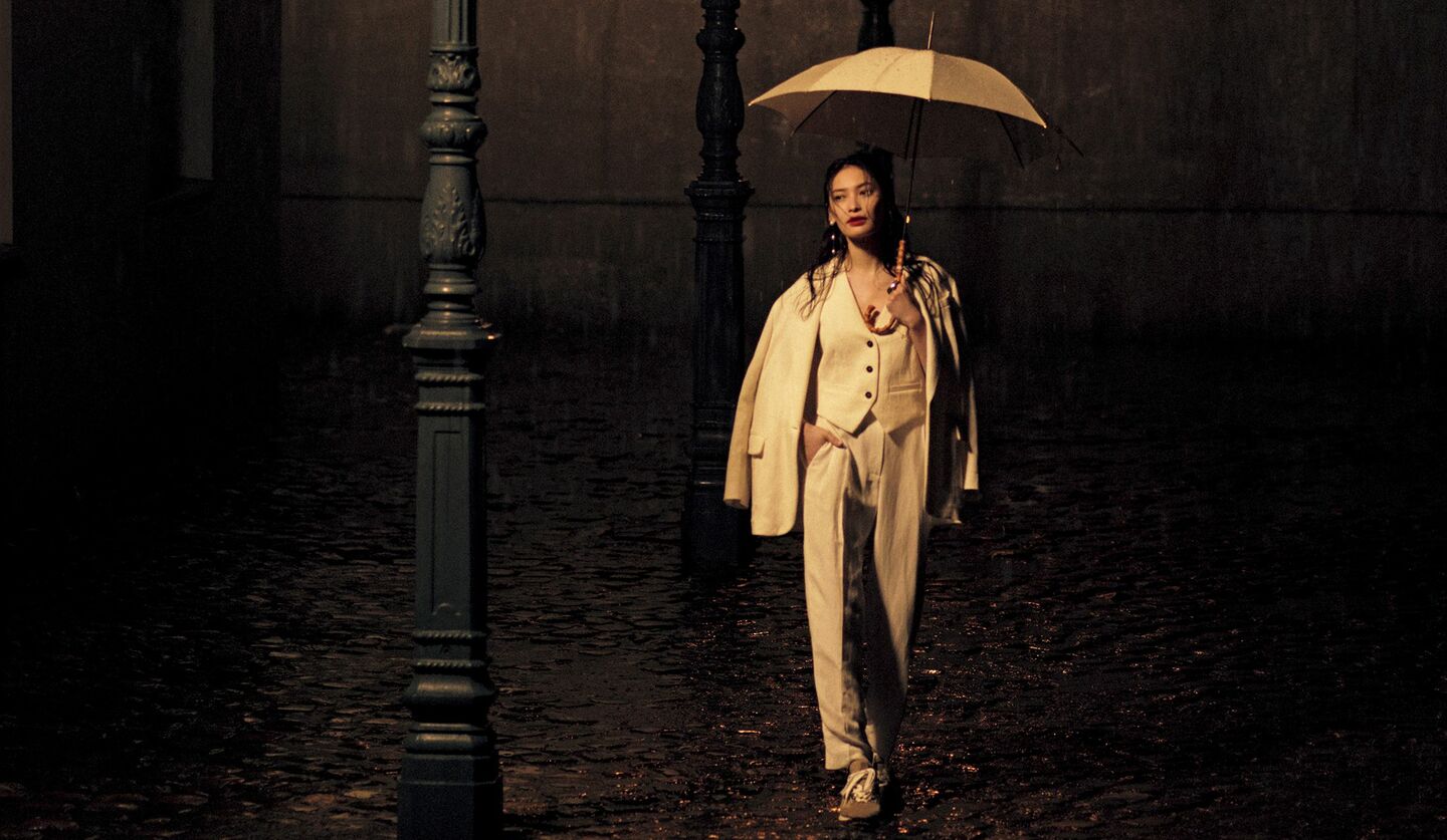ベージュのワントーンコーデで傘をさしながら町を歩くモデル大政絢さんの写真