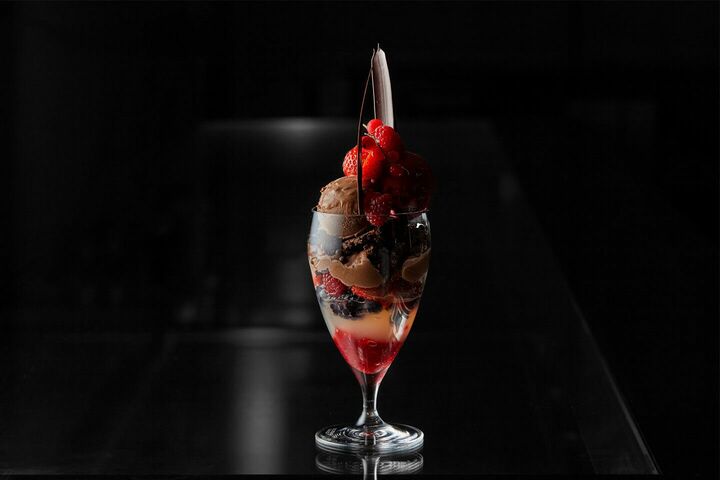 ワイングラスに赤いゼリー、白いゼリー、黒や紅のベリーやナッツ、チョコレートジェラート、ベリーのジェラートが層になり、ジェラートの上に真っ赤なベリーとカーブを描いたチョコレート板が飾られている