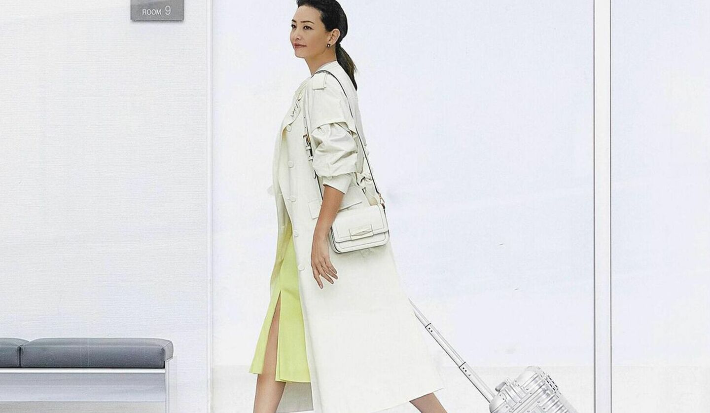 白のロングコートにイエローのスリットスカート、キャリーバックを持つモデルRINAさんの写真