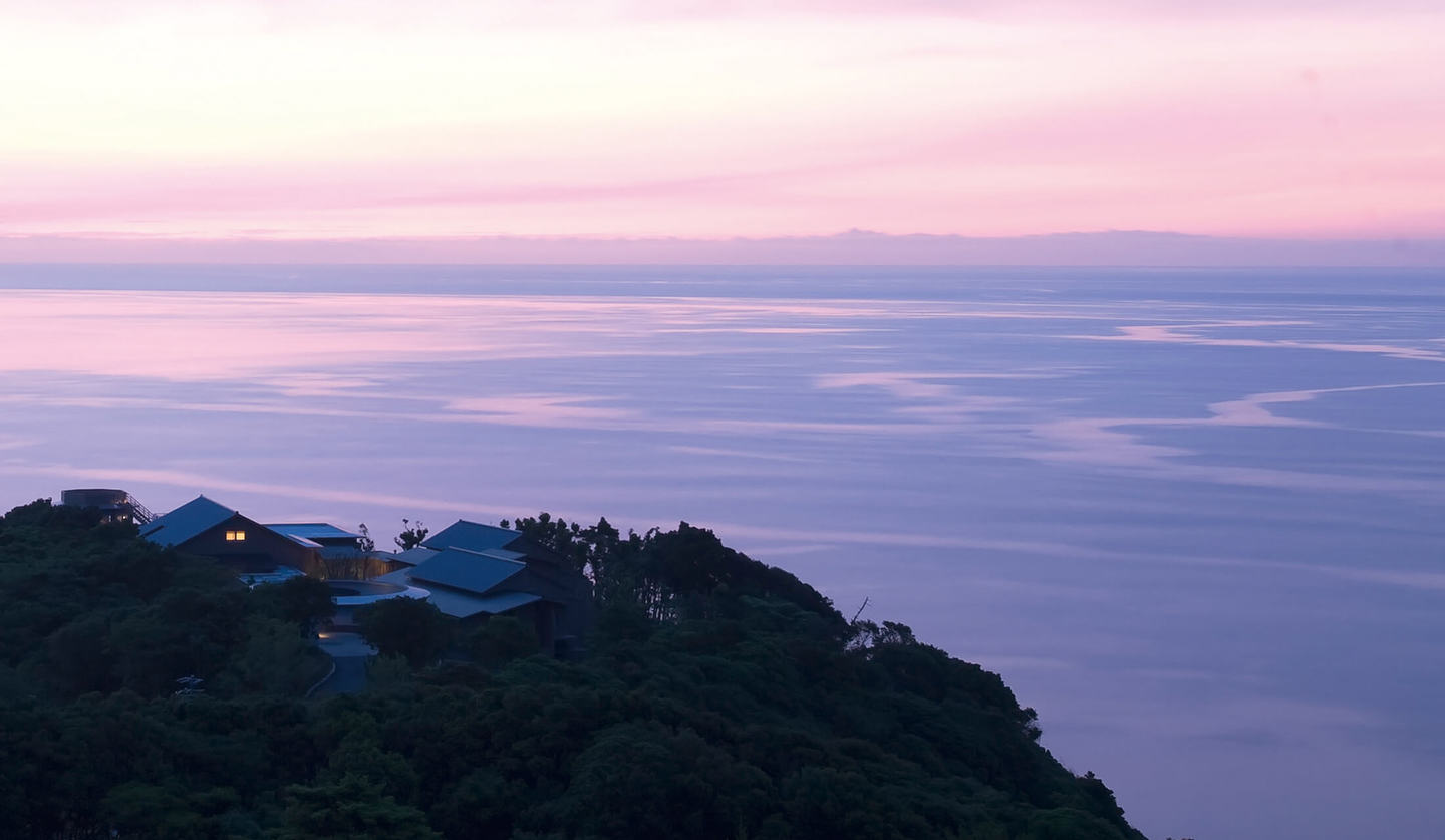 朝焼けで朱色にの空と赤紫になびく海にせり出した岬があり、岬の上に旅館が建っている